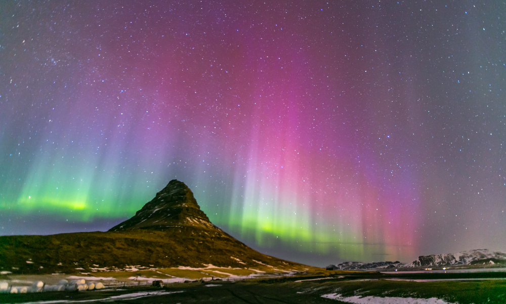 Image shows the Northern Lights illuminating a night sky with bands of green, purple, and pink light above a rocky landscape. A dark mountain rises in the foreground beneath a clear, star-filled sky