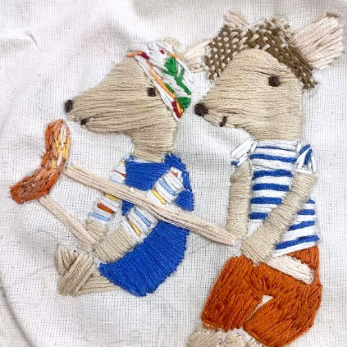 An embroidered textile artwork showing two stylised animals in clothing, stitched in thread on fabric with detailed textures and patterns.