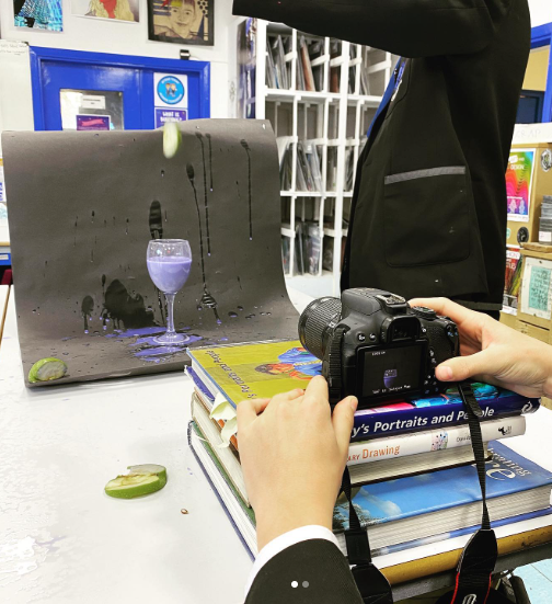 A pupil photographs a still-life setup using a digital camera balanced on art books, showing a glass, splashes of liquid, and fruit arranged against a dark backdrop in an art classroom.