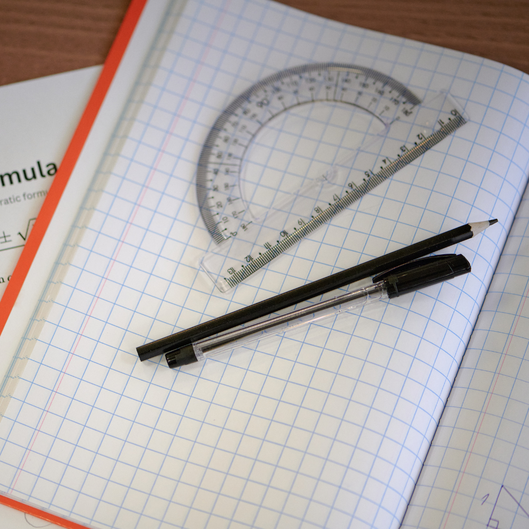 An open squared exercise book on a desk with a ruler, protractor, pen and pencil laid across the page