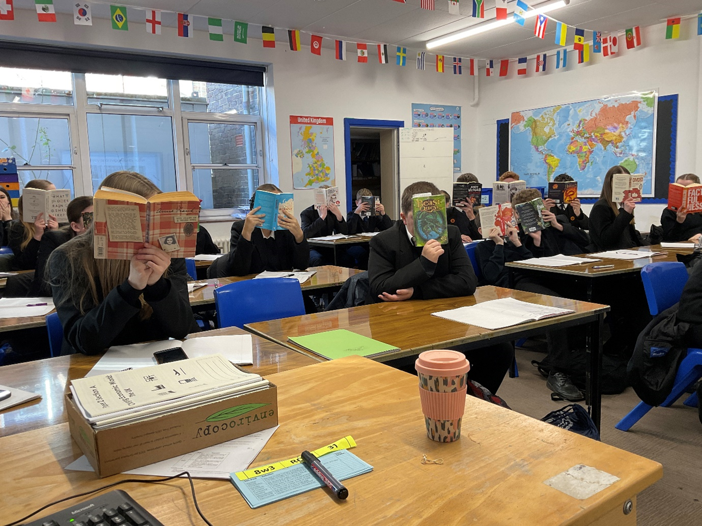 Image shows a classroom of secondary school pupils seated at desks, all holding books up and reading. The room is decorated with world maps and international flags, and exercise books, stationery, and classroom resources are visible on the desks.