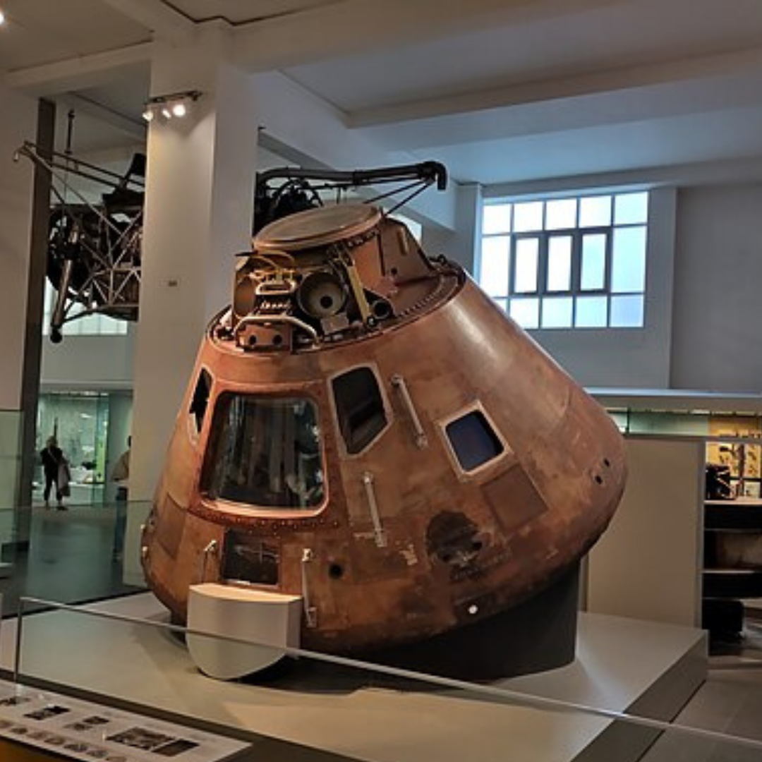Image shows the Apollo 10 Command Module displayed inside the Science Museum in London. The spacecraft is cone-shaped and covered in heat-scorched copper-brown metal, showing marks and wear from space travel and re-entry. Small square and trapezoid windows are visible around the upper section, along with panels, bolts, and technical components. The module is positioned indoors within a bright museum gallery, with white pillars, display cases, and large windows in the background.