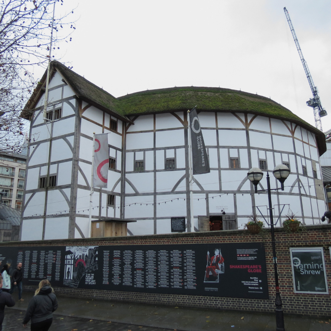 Image shows the exterior of Shakespeare’s Globe Theatre in London, a circular timber-framed building with a thatched roof, viewed from the street with banners displayed outside.