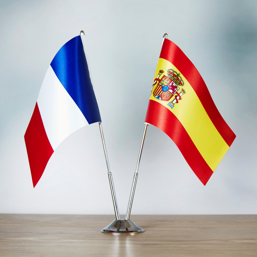 Two small desk flags, one representing France and one representing Spain, stand side-by-side in a silver chrome base on a wooden table against a soft blue and white background.
