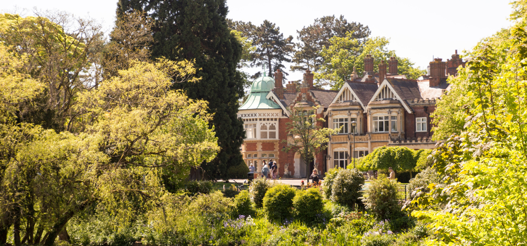 A view of Bletchley Park with historic red-brick buildings partially framed by trees and gardens, showing people walking along paths on a bright, sunny day.