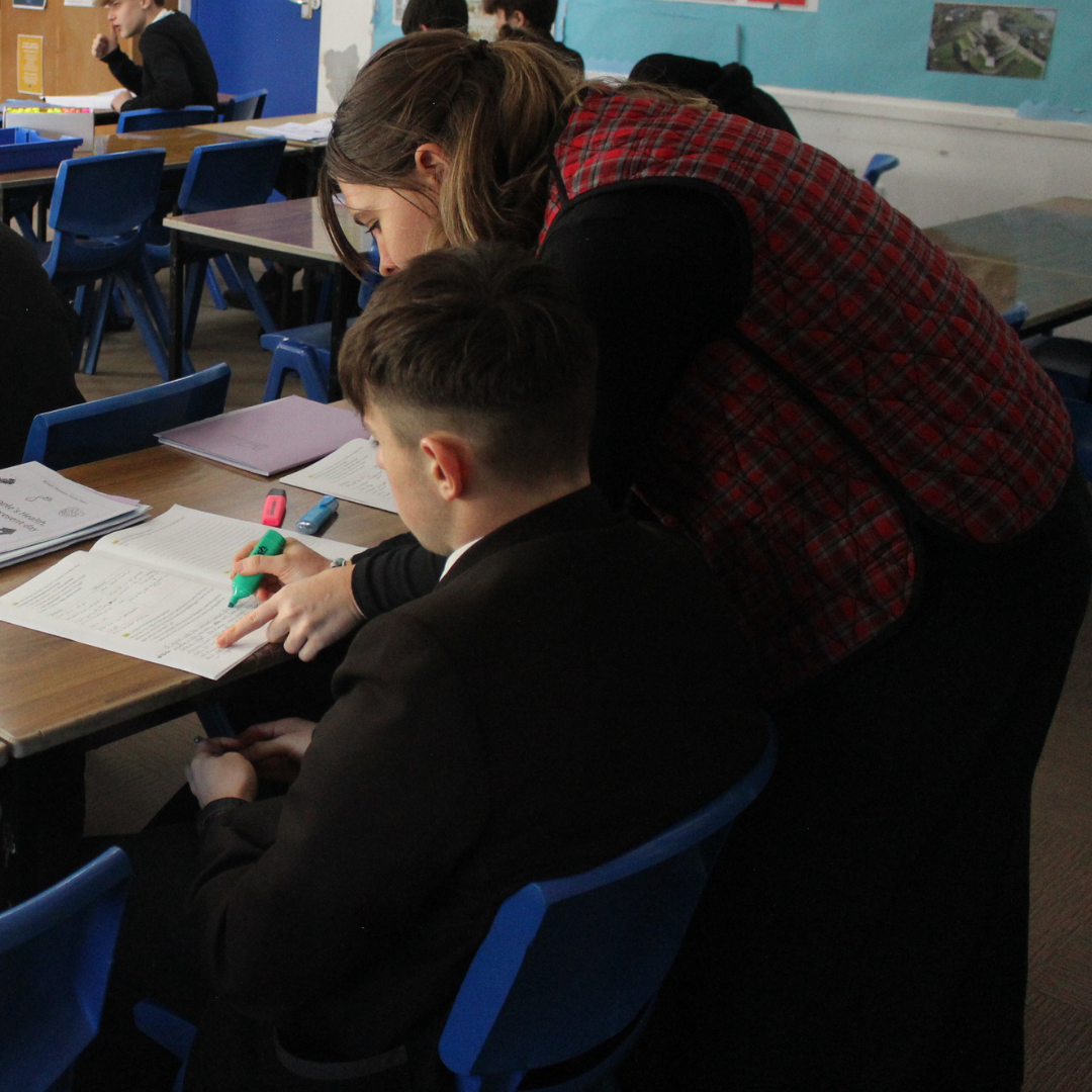 A close-up, candid shot of a teacher leaning over a desk to support a pupil during a lesson. The teacher is pointing to a workbook while the pupil uses a green highlighter to mark key information.