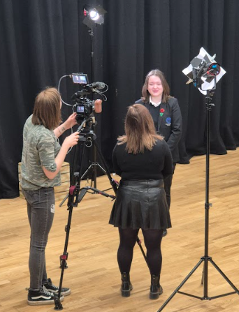 Image shows a pupil being filmed during a studio-style interview, with two pupils operating a camera and lighting equipment in a drama or media space.