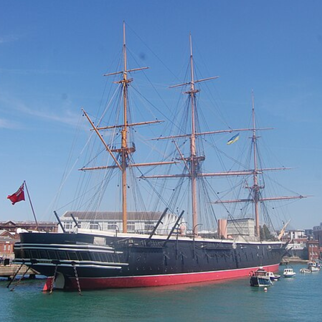 Image shows HMS Warrior moored at Portsmouth Historic Dockyard on a bright, clear day. The historic warship has a long black hull with tall wooden masts rising above it, crisscrossed with rigging and ropes. A red naval flag flies at the stern. Calm blue water is visible in the foreground, while dockyard buildings and modern structures can be seen behind the ship under a clear blue sky.