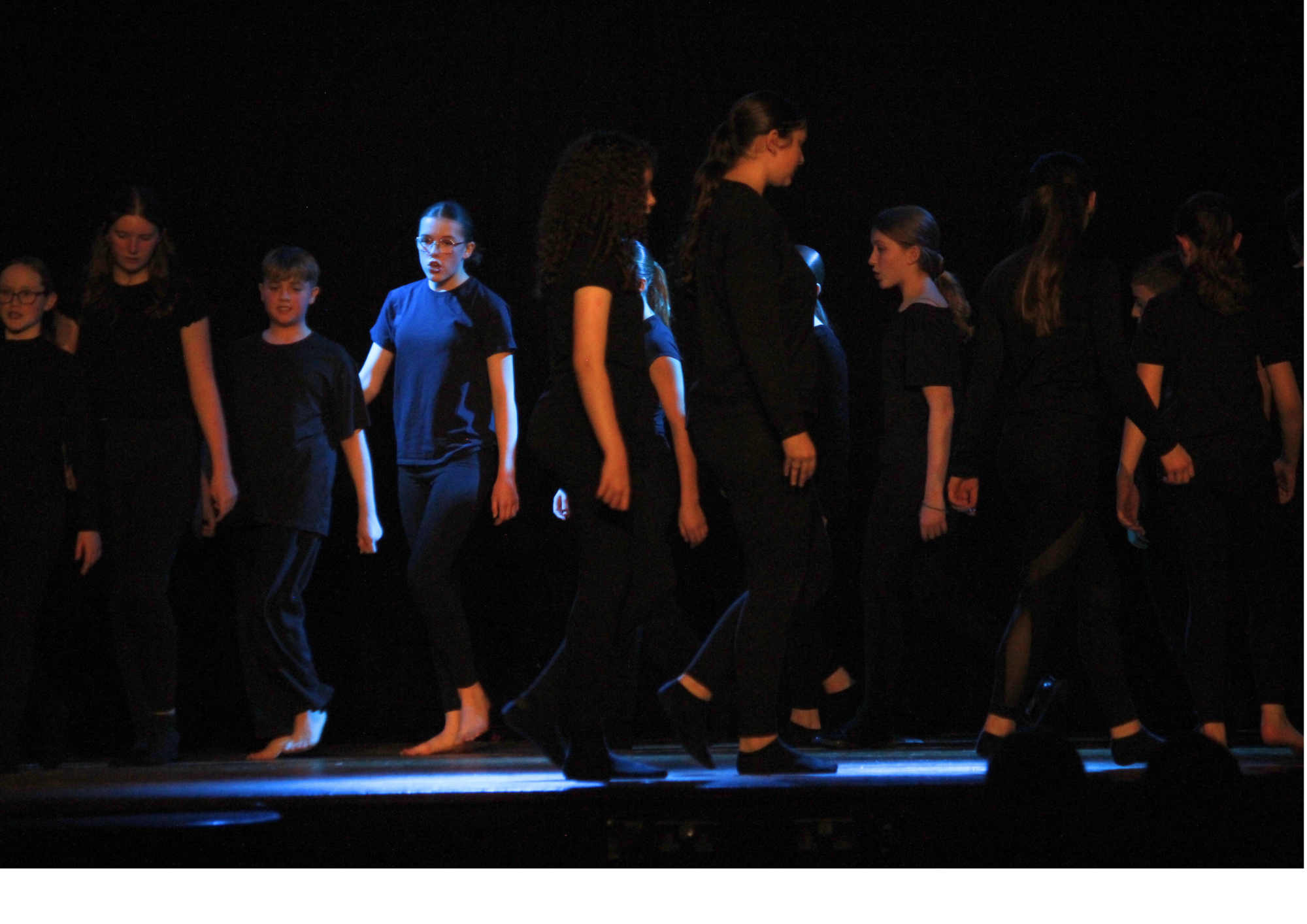Image shows a group of pupils dressed in black performing a drama or movement piece on a darkened stage, walking in coordinated lines under theatrical lighting.