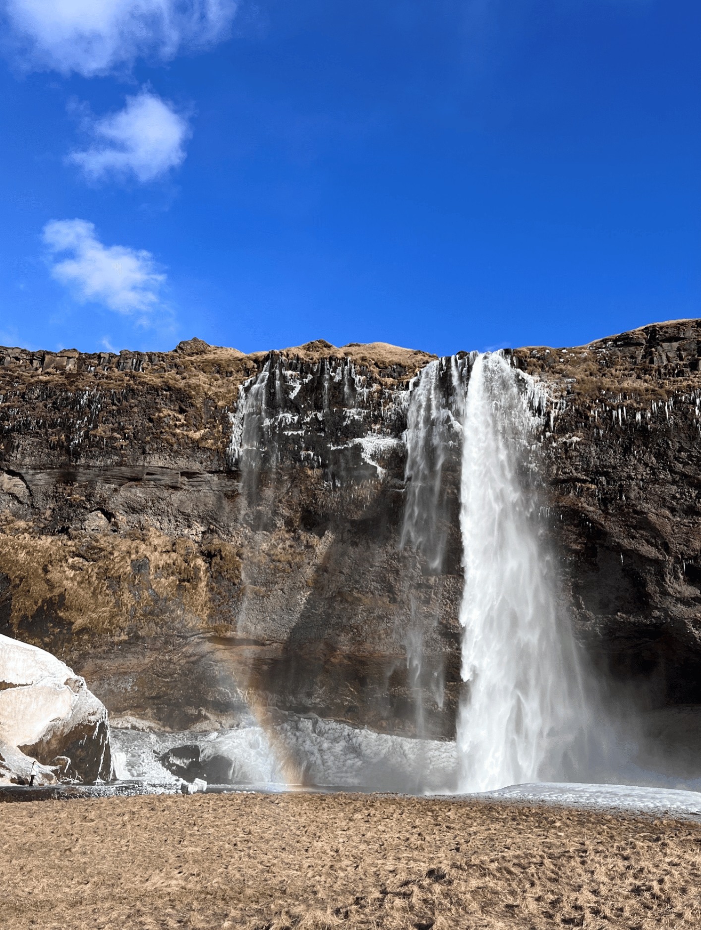 Image shows a tall waterfall cascading over a rocky cliff into a shallow pool below, with patches of ice at the base and a bright blue sky with scattered clouds above.