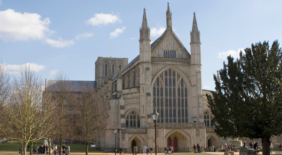 A wide daytime view of Winchester Cathedral, showing the large Gothic stone façade with tall arched windows and twin spires, set against a blue sky, with people walking across the cathedral close and trees framing the scene.
