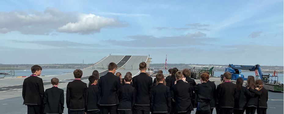 A rear-view shot of students in uniforms standing on the flight deck, observing the iconic ski-jump takeoff ramp extending toward the horizon under a light blue sky.