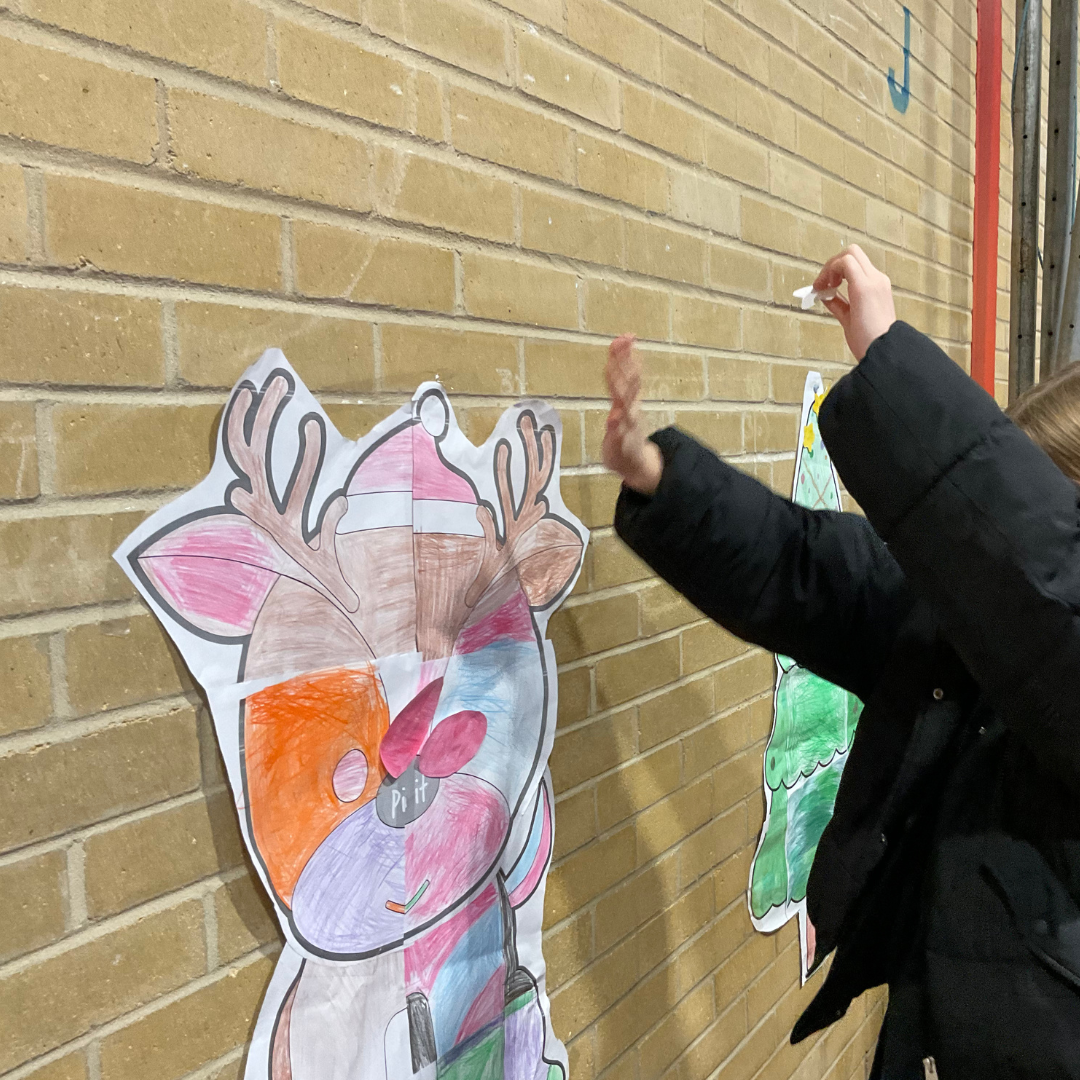 Image shows a coloured in Reindeer picture on the wall of the school hall. A pupil is trying to pin the red nose onto the reindeer. 