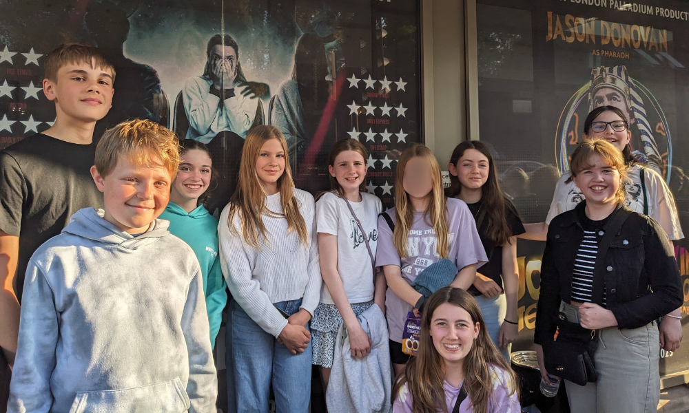 Image shows a group of ten pupils standing outside a theater in the sunshine. They are posed in front of large promotional posters for a West End production featuring Jason Donovan.