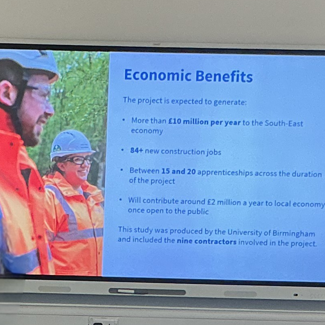 Close-up of a classroom display screen showing a presentation slide titled about economic benefits, with bullet points and figures alongside a photograph of construction workers wearing protective clothing and helmets.