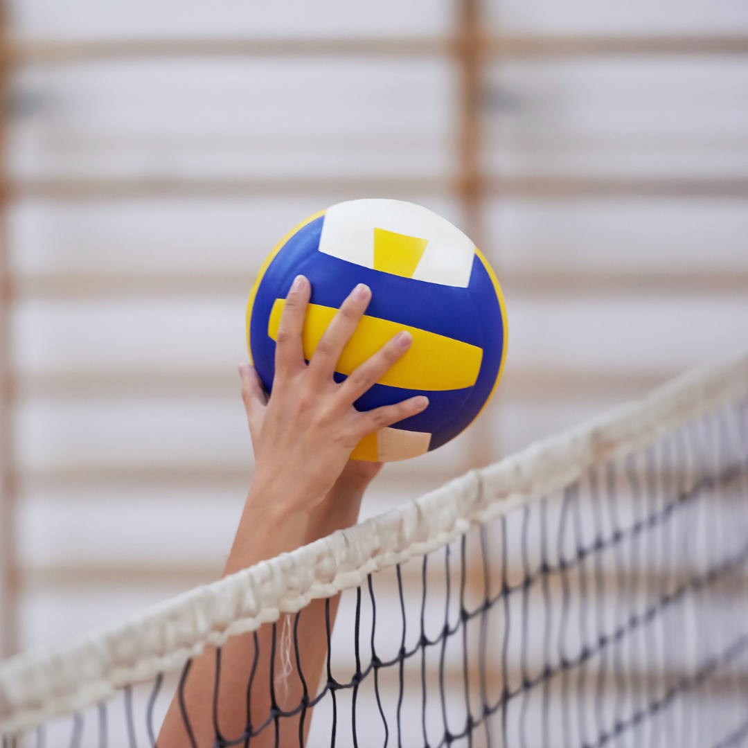 A close-up, slightly blurred action shot of a person’s hands reaching up to catch or block a blue, yellow, and white volleyball over a net in a sports hall.