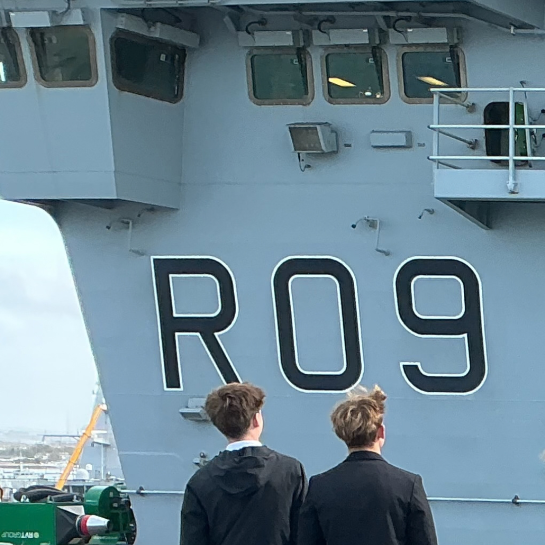 Two students in dark uniforms view the large white pennant identifier "R09" painted on the side of the aircraft carrier hull.