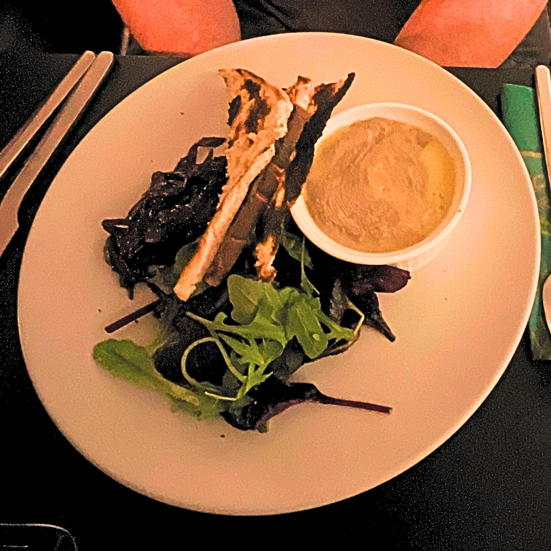 Starter plate with mixed salad leaves, grilled flatbread strips and a small ramekin of smooth pâté or dip on a white plate.