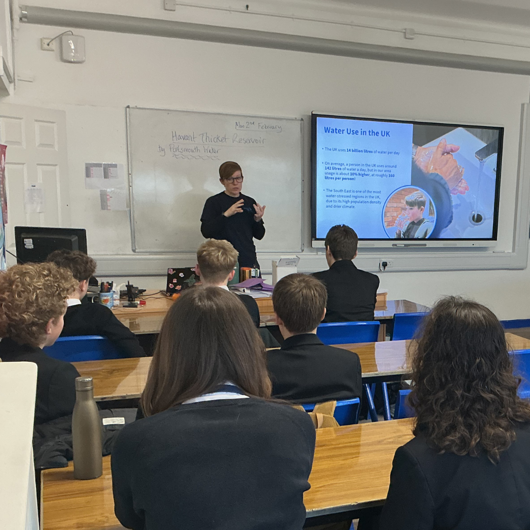 Adult presenter stands at the front of a classroom, gesturing while speaking to pupils seated at desks. A screen beside them shows a slide about water use in the UK, including images and bullet points, with a whiteboard visible behind.