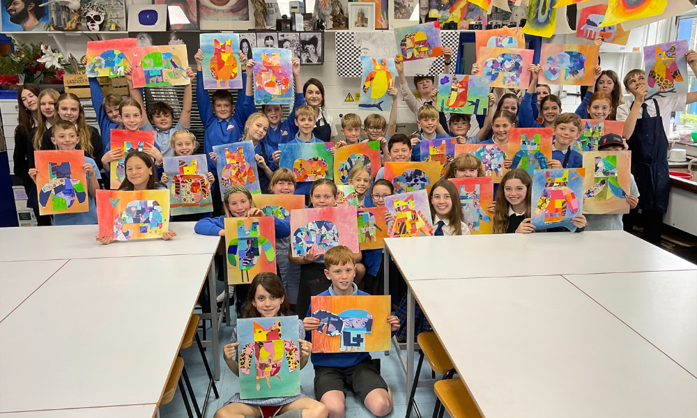 Image shows a large group of pupils in an art classroom, sitting and standing around white tables. Every pupil is smiling and holding up a vibrant, multi-colored collage of an animal (such as an elephant or owl) that they have created.