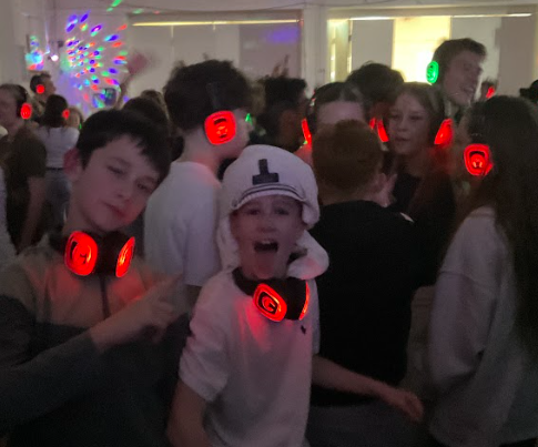 Image shows pupils dancing in a crowded school hall during a silent disco. Two boys in the foreground pose playfully for the camera while wearing glowing red headphones, with bright multicoloured disco lights projected onto the wall behind them.
