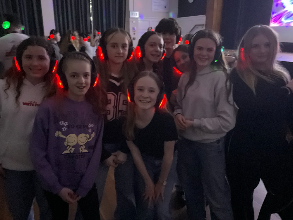 Image shows a group of smiling girls standing together at a silent disco in a school hall. They are wearing illuminated red headphones and casual clothing, posing for a photo while other pupils dance in the background under soft coloured lights.