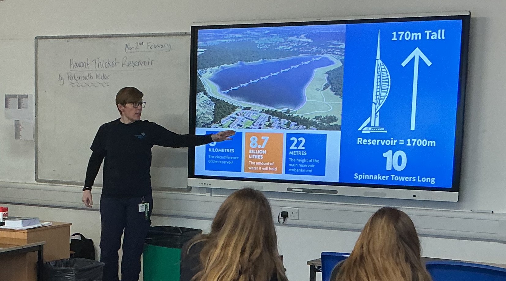 Wide view of a classroom with rows of pupils seated at desks, facing an adult presenter at the front. A large screen displays a slide related to the Havant Thicket Reservoir project, while classroom posters and resources are visible around the room.