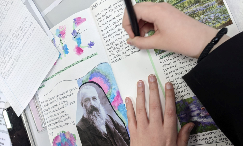 Image shows a close-up, top-down view of a pupil's hand using a black pen to write in an art sketchbook. The page features a research project on Claude Monet for the Bronze Arts Award, including a printed portrait of the artist, hand-painted colorful borders, and neat handwriting.