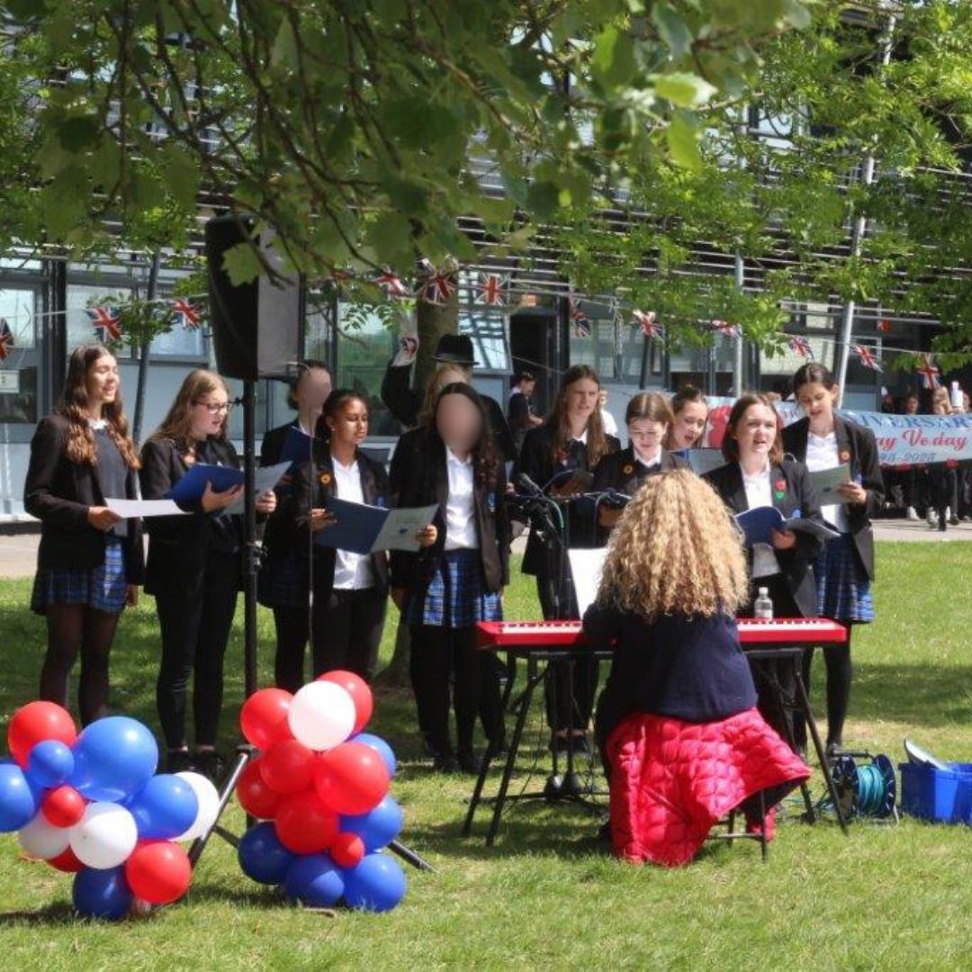 Image shows the College's Chamber Choir singing under the trees at the edge of the school field accompanied by Miss Shaw