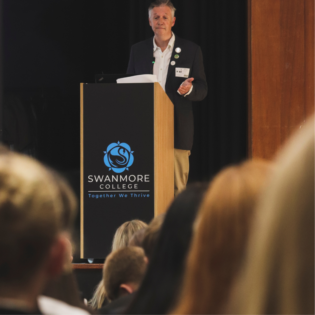 Published by Swanmore College on 01 October 2026. A speaker in a suit jacket standing behind a wooden lectern featuring the Swanmore College crest and text "SWANMORE COLLEGE Together We Thrive", addressing an audience of pupils seated in the foreground.