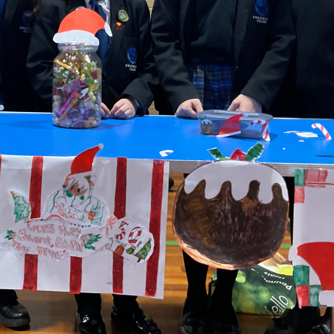 Image shows two pupils standing behind a stall decorated with a picture of a Christman pudding and a festive sign that reads "Guess the Sweets". There is a large jar of sweets on the stall with a paper Christmas hat on top.
