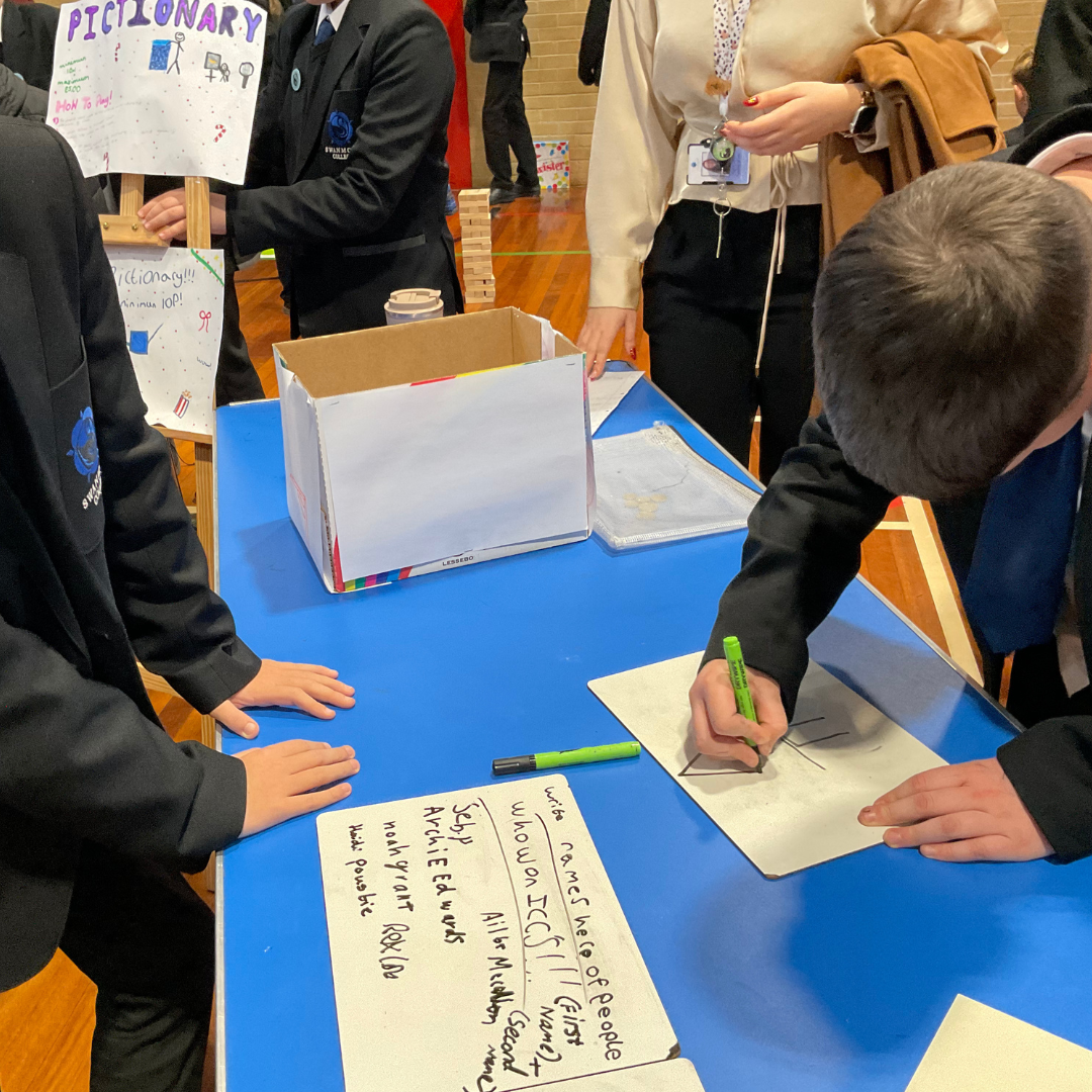 Image shows several pupils standing near a blue table in the school hall with one bending over the table and drawing. There is a white box on the table. There is also a member of staff standing next to the table. 