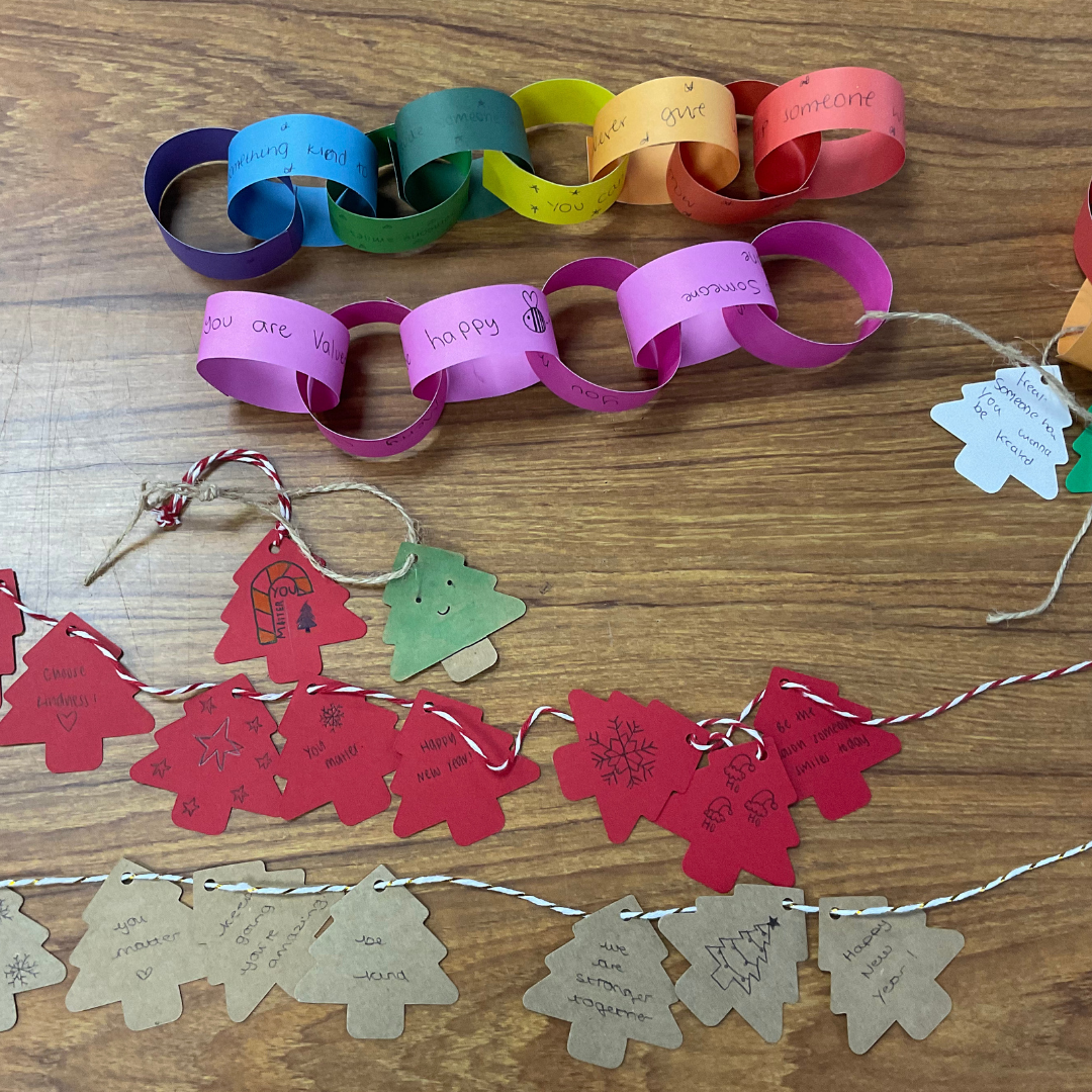 This image shows a rainbow paper link chain next to a string of Christmas tree decorations in red green and brown on a table