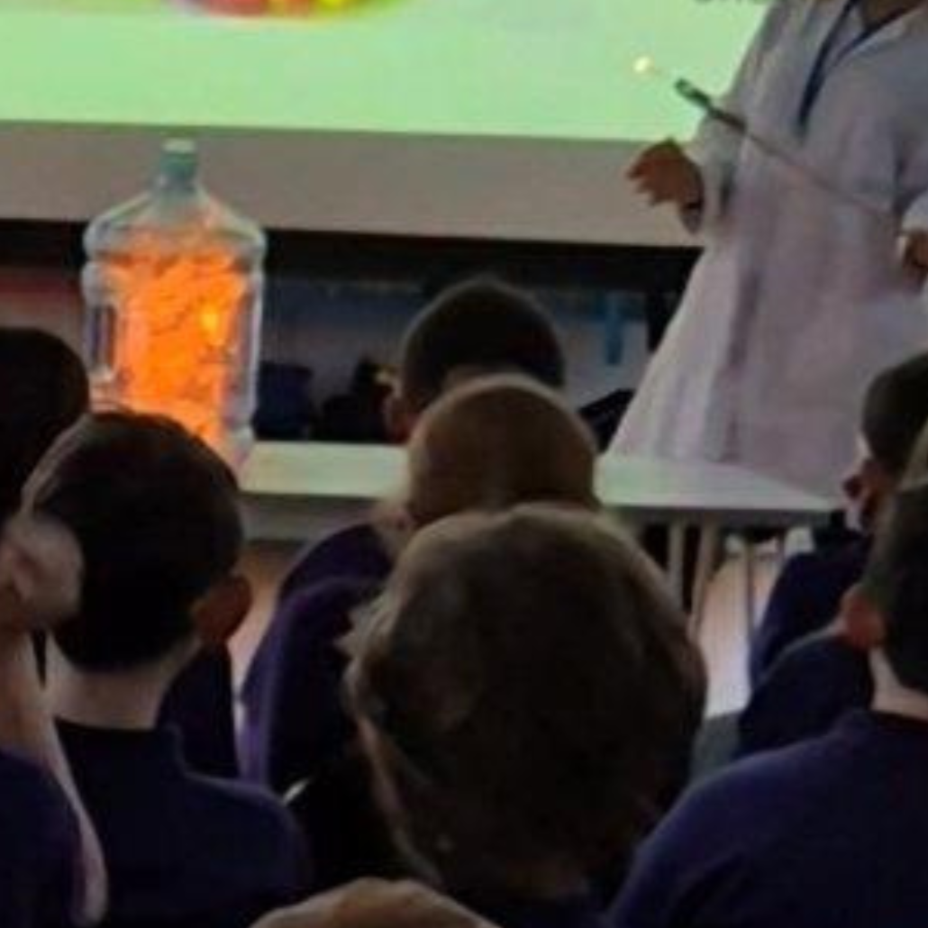 Bright orange flame filling the inside of a large plastic container during a controlled combustion demonstration in a primary school hall.