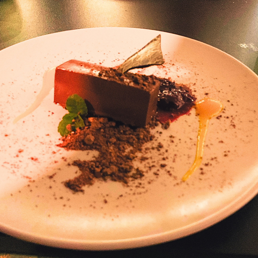 A plated dessert featuring a glossy rectangular mousse, chocolate crumb, fruit coulis, mint garnish and delicate chocolate shard on a white plate.