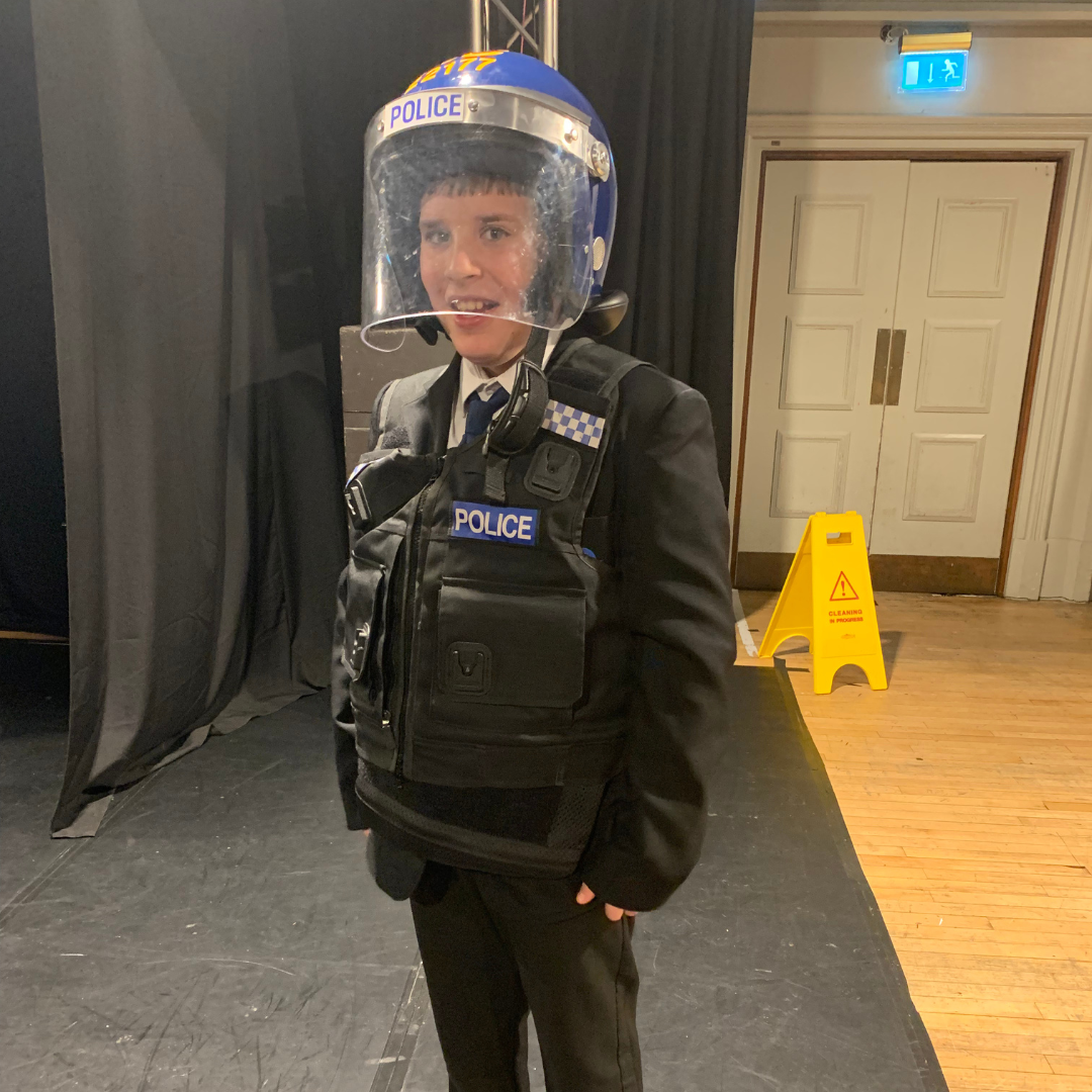 A pupil stands indoors wearing a full police riot gear uniform, including a heavy black tactical vest with "POLICE" written on the front and a blue helmet with a clear plastic visor. The pupil is smiling at the camera