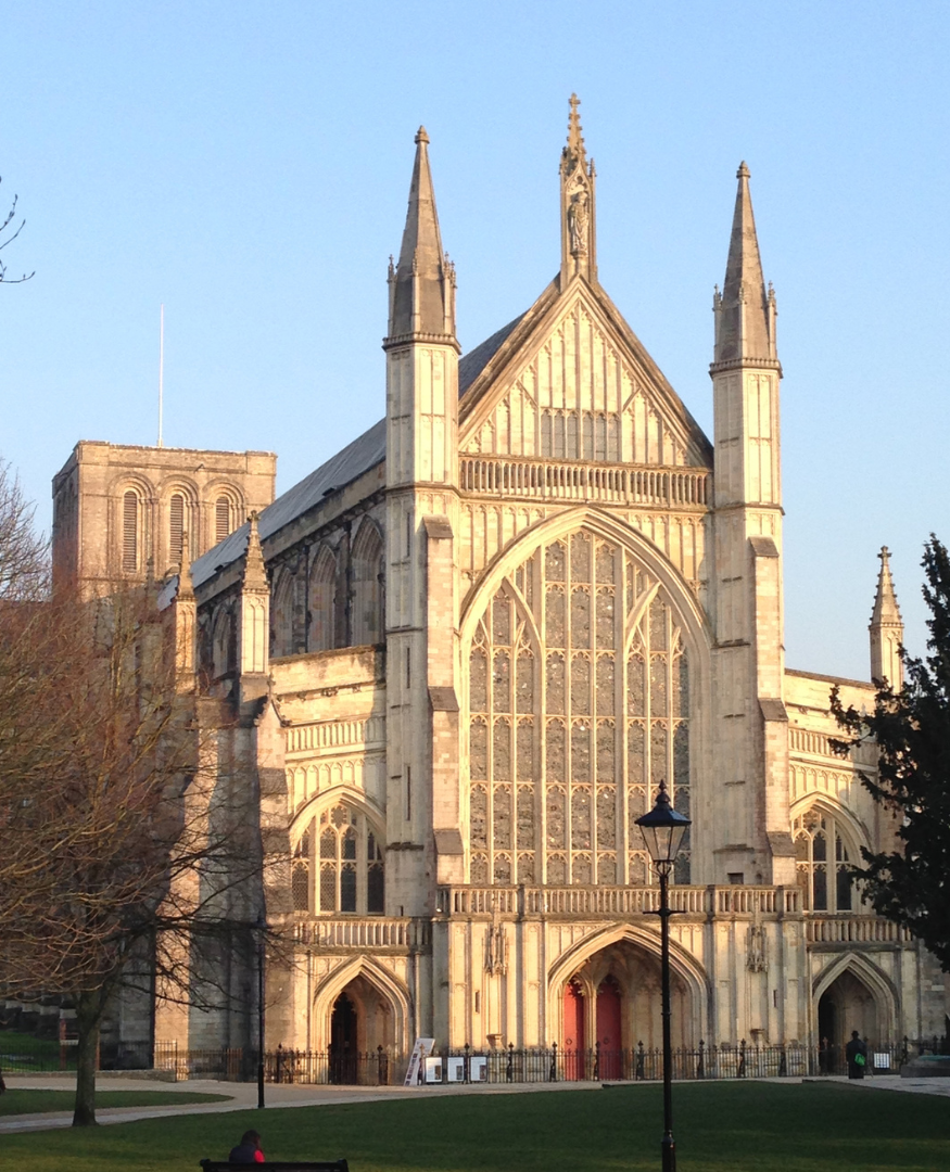 Exterior view of Winchester Cathedral, showing its large Gothic windows and towers in warm sunlight.