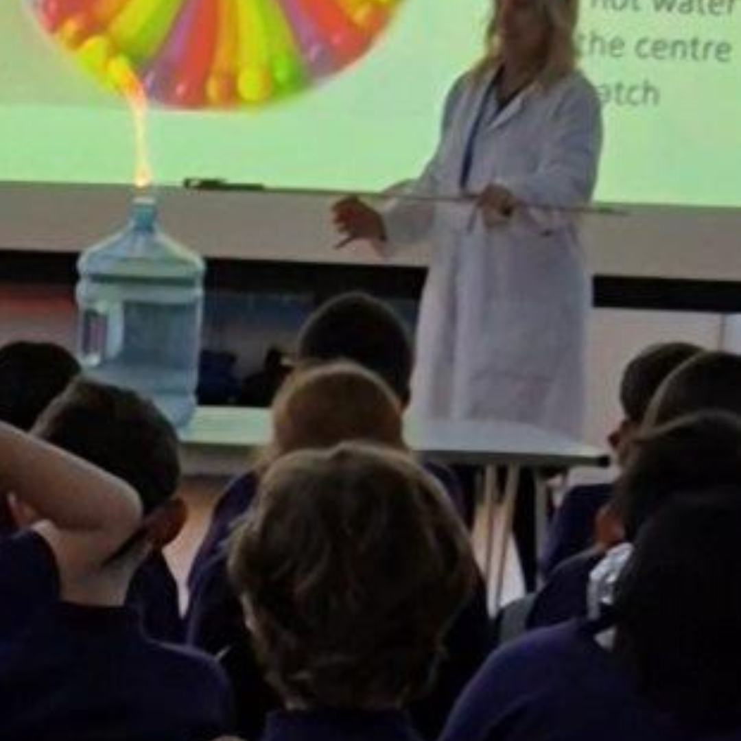 Science teacher in lab coat demonstrating a combustion experiment in a large water container, with a small flame visible at the top while primary pupils watch from the hall.