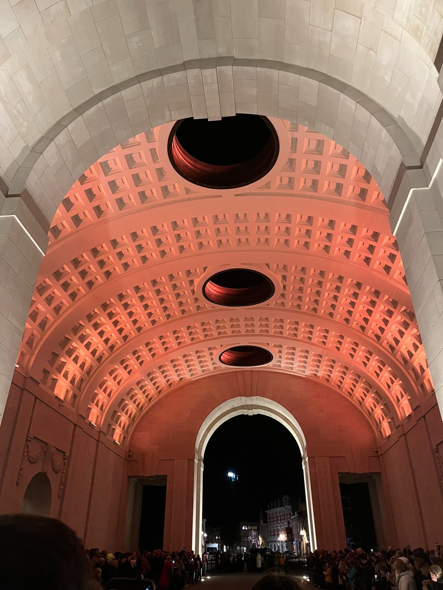 A view from inside the Menin Gate in Ypres at night, showing the illuminated memorial arches and crowd during the Last Post ceremony.