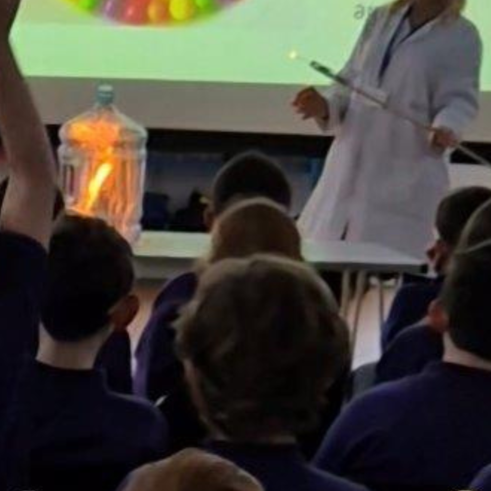 Combustion demonstration inside a large clear container with a visible burst of flame, pupils seated in front observing the experiment.
