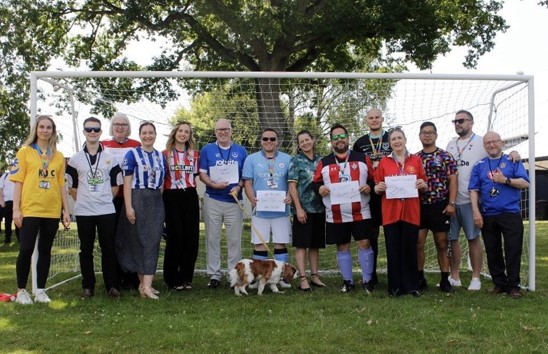 Staff members posing in football shirts beside a goal during a charity football-themed fundraising event.