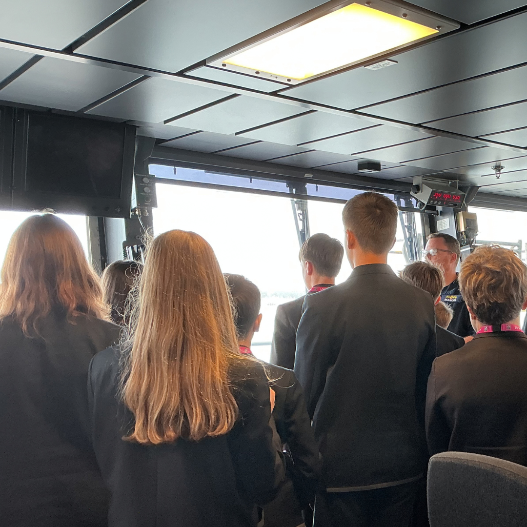 A group of students gathered inside the ship's bridge window area, listening to an instructor addressing them from the right.