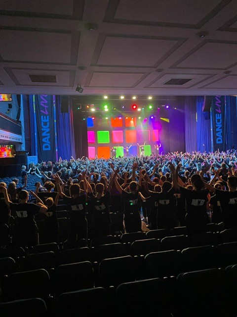 Image shows pupils in audience of theatre with presenters on stage at Dance Live rehearsal.
