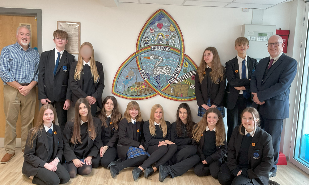 Image shows a group of thirteen pupils and two staff members posing in front of a large, colorful stained-glass style mosaic on a white wall. The mosaic is shaped like a triple-arch and features a swan and the words "Honesty," "Love," and "Respect."