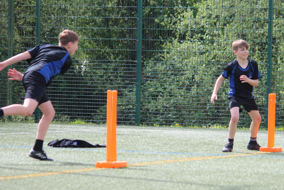 Two pupils in PE kit play cricket on an outdoor pitch, one bowling towards bright orange stumps while another prepares to field.