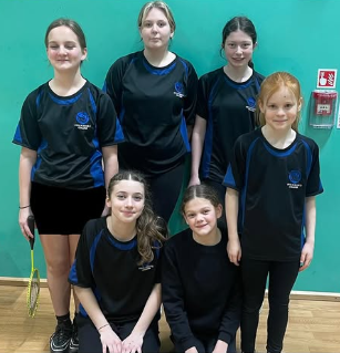 Six Key Stage 3 girls pose in a sports hall against a teal wall. They are wearing black and blue Swanmore College PE kit, with one pupil holding a badminton rackets.