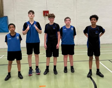 Five Year 9 boys stand side by side in a sports hall wearing blue and black Swanmore College basketball kit and black shorts. They are positioned on an indoor court with a white brick wall and red fire alarm behind them.