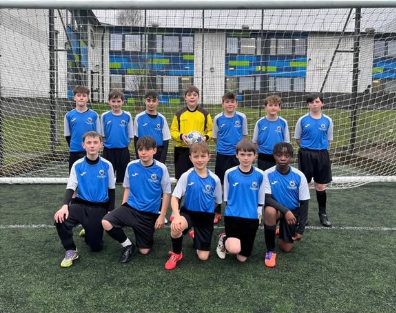 Group photo of 11 Year 8 boys in blue Swanmore College football kits posing in front of a goal on an outdoor 3G pitch. One pupil stands in the back row wearing a yellow goalkeeper top. A school building is visible behind the net.