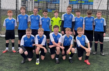 The Year 8 boys football squad pose for a team photo on an outdoor pitch in front of a goal. Some pupils kneel at the front while others stand behind. They are wearing blue and white football shirts with black shorts, with a goalkeeper in a bright yellow top.