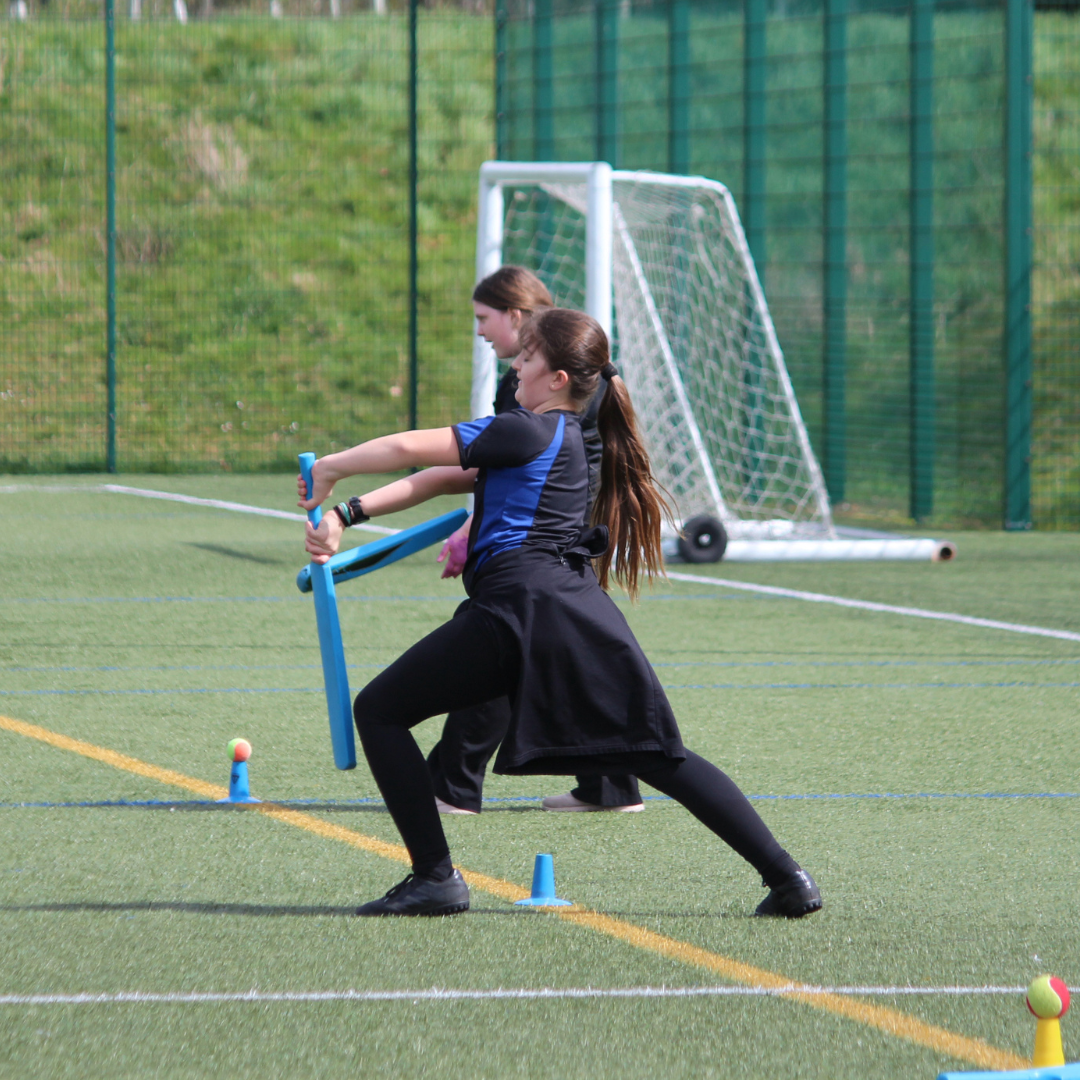 Two pupils practise batting with plastic cricket equipment on the school field, focusing on striking the ball during a drill.